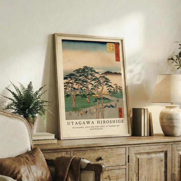May include: Framed Japanese woodblock print reproduction featuring Mount Fuji in background with traditional pine trees, small figures, and mist-covered landscape in muted greens, blues and earth tones. Light wood frame displayed on rustic wooden sideboard alongside fern plant, ceramic vase, and stacked books in neutral-toned living room setting.