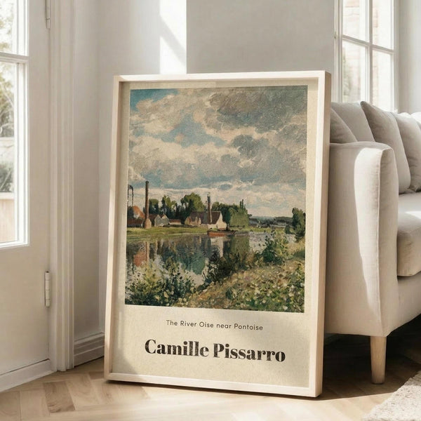May include: Framed art print leaning against white wall next to beige sofa in bright living room. Print features impressionist landscape painting with river, village buildings, and cloudy sky in blues, greens, and earth tones. Text below artwork reads "The River Oise near Pontoise" and "Camille Pissarro" on cream background. Light wood frame, hardwood floors visible.