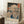 May include: Framed Japanese woodblock print showing snowy mountain landscape with traditional buildings, pine trees, and blue water. Print features muted earth tones with Japanese text characters in red cartouches. Modern light wood frame leaning against white striped wallpaper near wooden side table with white coffee cup and cream throw blanket on textured white rug.