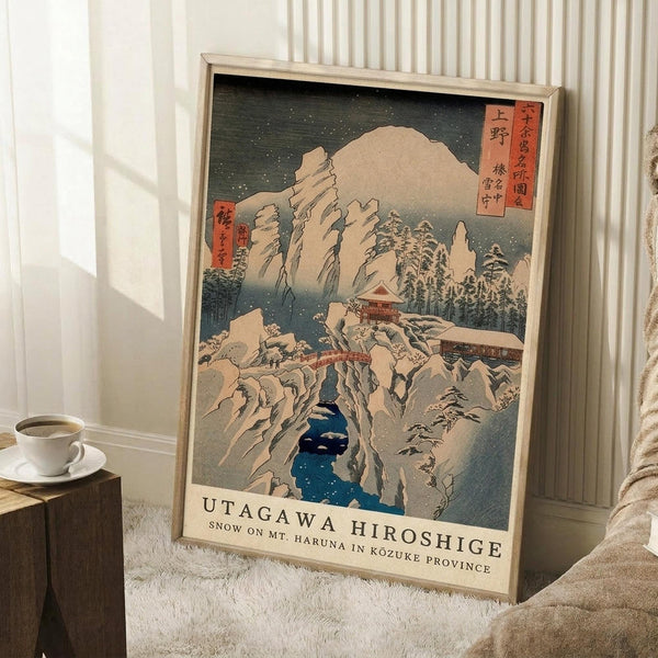 May include: Framed Japanese woodblock print showing snowy mountain landscape with traditional buildings, pine trees, and blue water. Print features muted earth tones with Japanese text characters in red cartouches. Modern light wood frame leaning against white striped wallpaper near wooden side table with white coffee cup and cream throw blanket on textured white rug.
