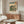 May include: Japanese woodblock print artwork displayed in modern living room with wooden sideboard, white curved chair, glass coffee table, and floor lamp. The vintage-style poster shows a landscape scene with Mount Fuji, green trees, and figures in traditional dress, featuring soft blue, green and orange tones in an ornate frame against neutral walls.