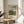 May include: Framed art print featuring an impressionist landscape painting with cloudy blue sky, green trees, and buildings reflected in water. Print has cream matting with "Camille Pissarro" text below the artwork. Thin gold frame sits on round wooden side table next to beige upholstered chair. Soft natural lighting creates shadows on white wall. Palm plant leaves visible in corner.