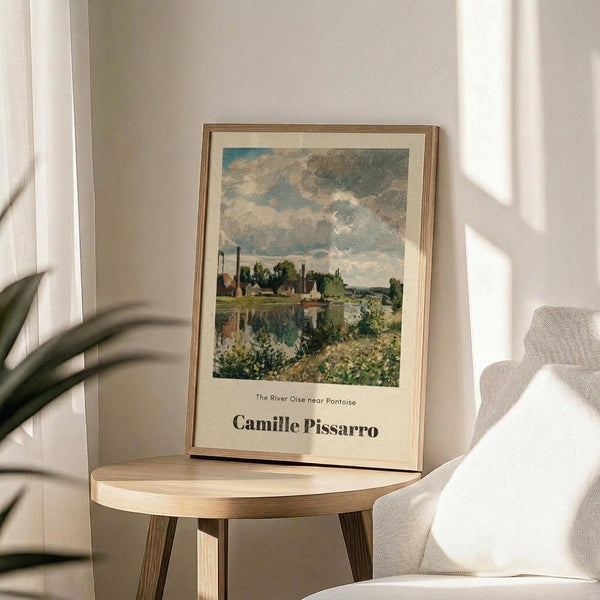 May include: Framed art print featuring an impressionist landscape painting with cloudy blue sky, green trees, and buildings reflected in water. Print has cream matting with "Camille Pissarro" text below the artwork. Thin gold frame sits on round wooden side table next to beige upholstered chair. Soft natural lighting creates shadows on white wall. Palm plant leaves visible in corner.