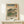 May include: Framed Japanese woodblock print reproduction featuring Mount Fuji in the background with pine trees, green landscape, and small figures along a path. Print has vintage beige tones with Japanese text and red cartouche. Displayed on light wood surface with small white wrapped gift beside natural wood frame.