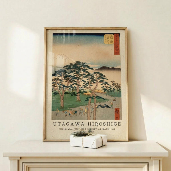 May include: Framed Japanese woodblock print reproduction featuring Mount Fuji in the background with pine trees, green landscape, and small figures along a path. Print has vintage beige tones with Japanese text and red cartouche. Displayed on light wood surface with small white wrapped gift beside natural wood frame.