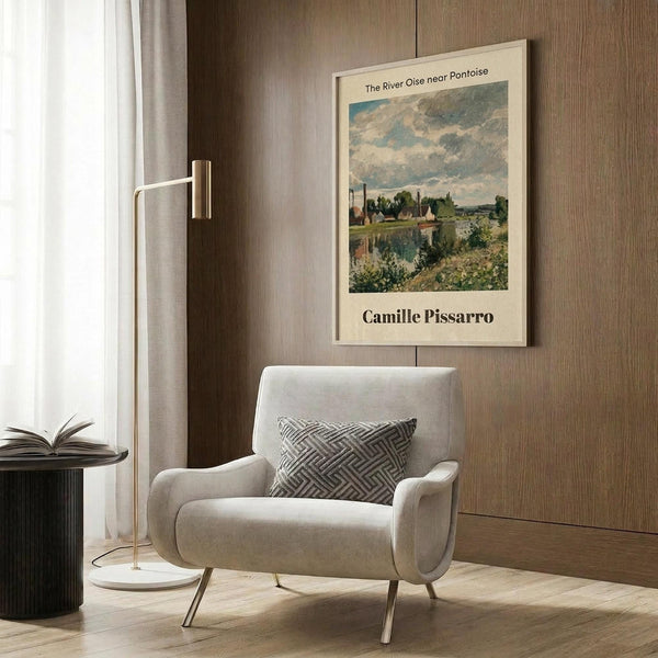 May include: Beige framed art print featuring a French countryside landscape with river, buildings, and cloudy sky in impressionist style, displayed on wood-paneled wall above cream upholstered armchair with geometric patterned pillow, brass floor lamp, and dark round side table with open book in modern living room setting.