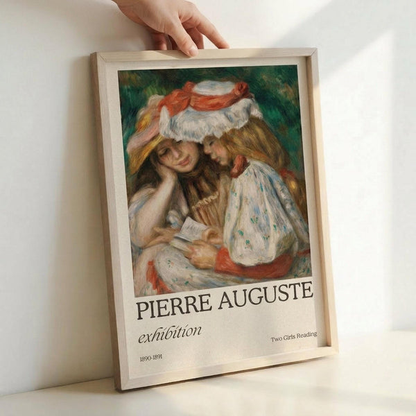 May include: Hand placing framed art poster against white wall. The poster features a soft, impressionist-style painting of two young women with long hair reading together, one wearing a white hat with red ribbon. Text below reads "PIERRE AUGUSTE exhibition" with "Two Girls Reading" and "1890-1891" in smaller print. Light wooden frame surrounds the cream-colored poster with painterly brushstrokes in warm tones.