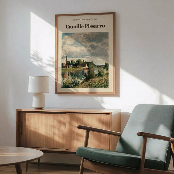 May include: Framed art print of impressionist river landscape with boats, trees and cloudy sky in soft blues and greens, displayed above wood credenza with ceramic lamp, next to mid-century modern sage green upholstered chair with walnut arms in bright sunlit room with white walls