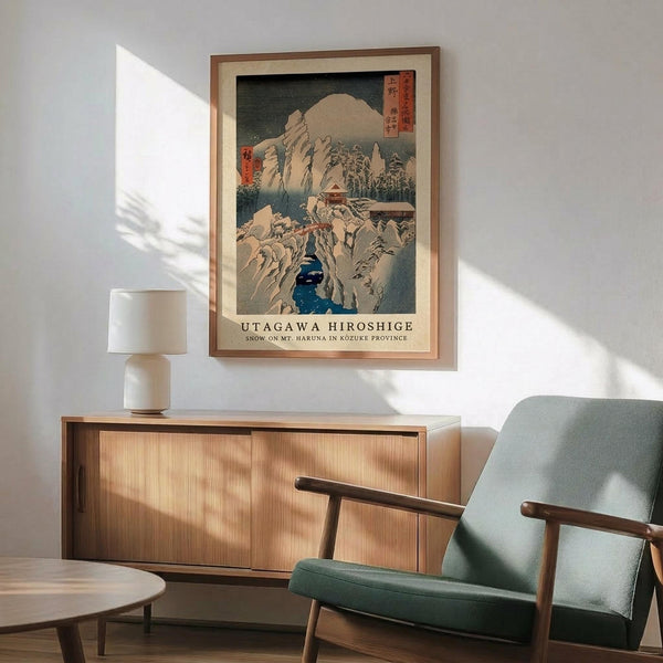 May include: Japanese woodblock print poster in wooden frame showing snow-covered mountains with blue river gorge and traditional buildings. Displayed on white wall above natural wood sideboard with ceramic table lamp. Mid-century modern green upholstered chair with wooden arms positioned nearby. Warm sunlight creates geometric shadows across the minimalist room setting.