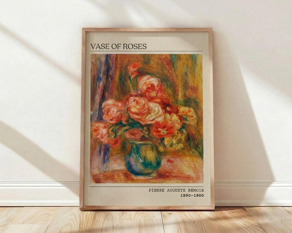 May include: Framed poster print of Renoir's impressionist roses painting leaning against white wall on light wood floor. Artwork shows pink and coral roses in blue-green vase with warm yellow-orange background brushstrokes. Cream matting surrounds the colorful floral image, with artist name and dates printed below. Natural window light creates soft shadows on wooden frame and floor.