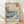 May include: Framed poster print of Japanese woodblock art featuring Mount Fuji in background, large blue ocean waves in foreground, sailing boat, and flying birds. The artwork displays traditional ukiyo-e style with blue, white, and earth tones. Red text cartouches visible in corners. Frame appears light wood, leaning against white wall on wooden floor with natural lighting creating soft shadows.