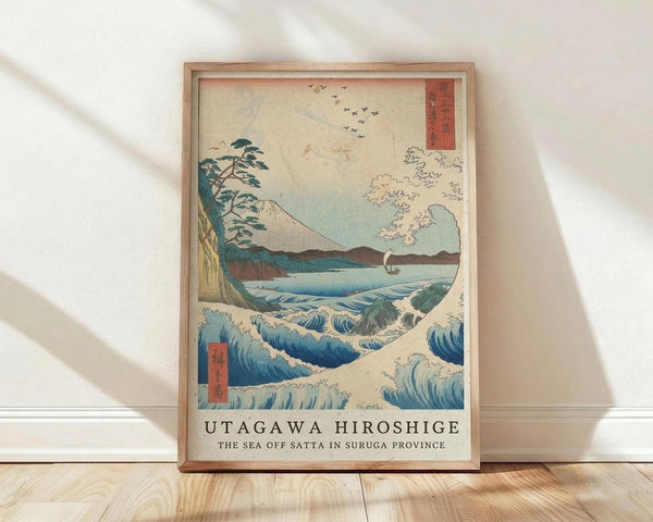 May include: Framed poster print of Japanese woodblock art featuring Mount Fuji in background, large blue ocean waves in foreground, sailing boat, and flying birds. The artwork displays traditional ukiyo-e style with blue, white, and earth tones. Red text cartouches visible in corners. Frame appears light wood, leaning against white wall on wooden floor with natural lighting creating soft shadows.