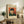 May include: Framed Van Gogh self-portrait poster on wooden console table in living room setting. Shows the artist wearing a fur hat and green coat, smoking a pipe against orange-red background. Cream-colored matting and thin wooden frame. Displayed alongside fern plant, ceramic vase, books, and brown leather chair visible in cozy, neutral-toned interior.