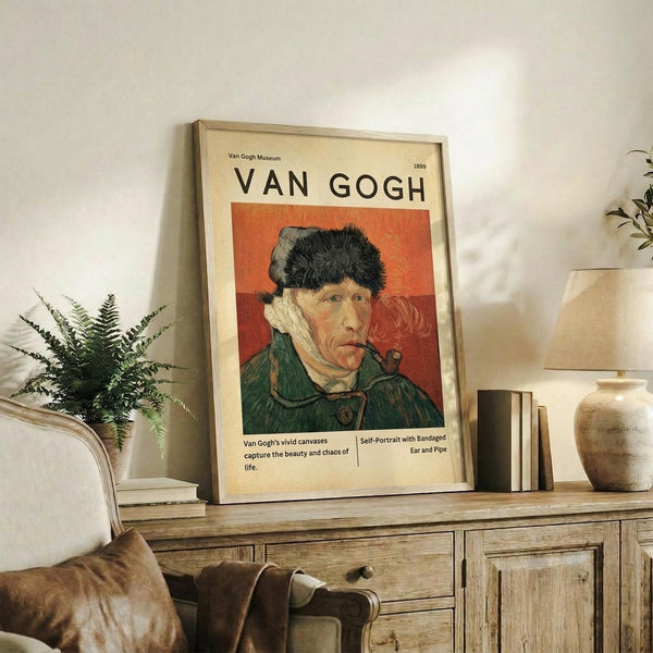 May include: Framed Van Gogh self-portrait poster on wooden console table in living room setting. Shows the artist wearing a fur hat and green coat, smoking a pipe against orange-red background. Cream-colored matting and thin wooden frame. Displayed alongside fern plant, ceramic vase, books, and brown leather chair visible in cozy, neutral-toned interior.