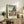 May include: Framed Japanese woodblock print reproduction displayed on rustic wooden sideboard in neutral-toned living room. Artwork shows coastal landscape with rocky cliffs, blue water, and traditional Japanese text in coral and blue tones. Scene includes fern plant, ceramic vase, stacked books, and beige upholstered chair with brown leather pillow.