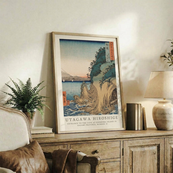 May include: Framed Japanese woodblock print reproduction displayed on rustic wooden sideboard in neutral-toned living room. Artwork shows coastal landscape with rocky cliffs, blue water, and traditional Japanese text in coral and blue tones. Scene includes fern plant, ceramic vase, stacked books, and beige upholstered chair with brown leather pillow.