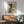 May include: Framed vintage Japanese woodblock print showing cascading waterfall with wooden bridge, displayed above mid-century modern wooden sideboard with white table lamp. Sage green upholstered armchair with wooden frame sits nearby on wooden floor. Natural sunlight creates geometric shadows on white wall. Print features muted blues, greens, and earth tones in traditional artistic style.