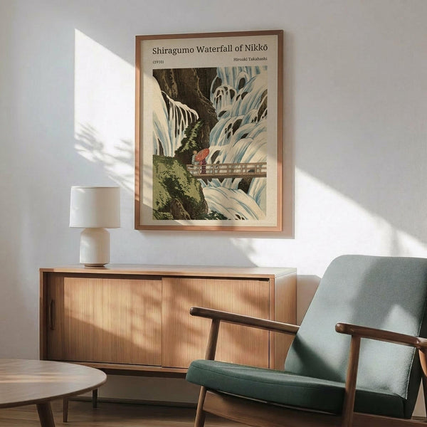 May include: Framed vintage Japanese woodblock print showing cascading waterfall with wooden bridge, displayed above mid-century modern wooden sideboard with white table lamp. Sage green upholstered armchair with wooden frame sits nearby on wooden floor. Natural sunlight creates geometric shadows on white wall. Print features muted blues, greens, and earth tones in traditional artistic style.