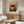 May include: Van Gogh museum poster featuring self-portrait of artist wearing dark fur hat and green coat against orange background, displayed above wooden sideboard in modern living room with white upholstered chair, glass coffee table, and minimalist decor in neutral tones