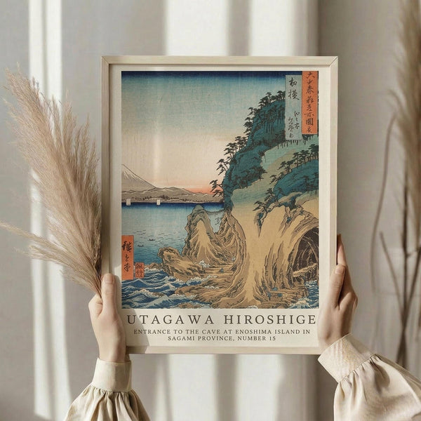 May include: Person holding framed Japanese woodblock print showing coastal cliff scene with cave entrance, rocky formations, blue water, and forested hills. Print features traditional ukiyo-e style with soft blues, greens, and earth tones, displayed in light wood frame. Person wears cream sweater, holds dried pampas grass, standing against white curtained background.