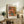 May include: Framed reproduction of Renoir's impressionist roses painting displayed on rustic wooden sideboard alongside fern plant, stacked books, and ceramic vases. Artwork shows pink and coral roses in blue-green vase with warm, painterly brushstrokes. Natural lighting creates soft shadows on white wall in cozy living room setting with beige upholstered furniture.