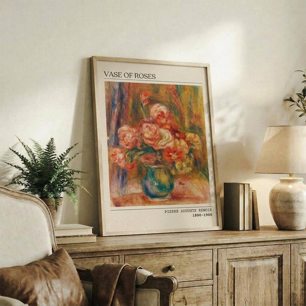 May include: Framed reproduction of Renoir's impressionist roses painting displayed on rustic wooden sideboard alongside fern plant, stacked books, and ceramic vases. Artwork shows pink and coral roses in blue-green vase with warm, painterly brushstrokes. Natural lighting creates soft shadows on white wall in cozy living room setting with beige upholstered furniture.