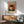 May include: Van Gogh self-portrait poster with bandaged ear and pipe, displayed above mid-century modern wooden credenza with table lamp. Sage green upholstered armchair with wooden frame sits nearby on wooden floor. Warm sunlight creates shadows across white wall in contemporary living room setting.