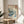 May include: Framed reproduction of Japanese woodblock print showing dramatic ocean waves, Mount Fuji, flying birds, and Japanese text, displayed on round wooden side table next to beige upholstered chair with plant leaves visible in bright, naturally-lit room with white walls.