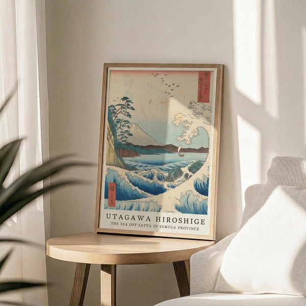 May include: Framed reproduction of Japanese woodblock print showing dramatic ocean waves, Mount Fuji, flying birds, and Japanese text, displayed on round wooden side table next to beige upholstered chair with plant leaves visible in bright, naturally-lit room with white walls.