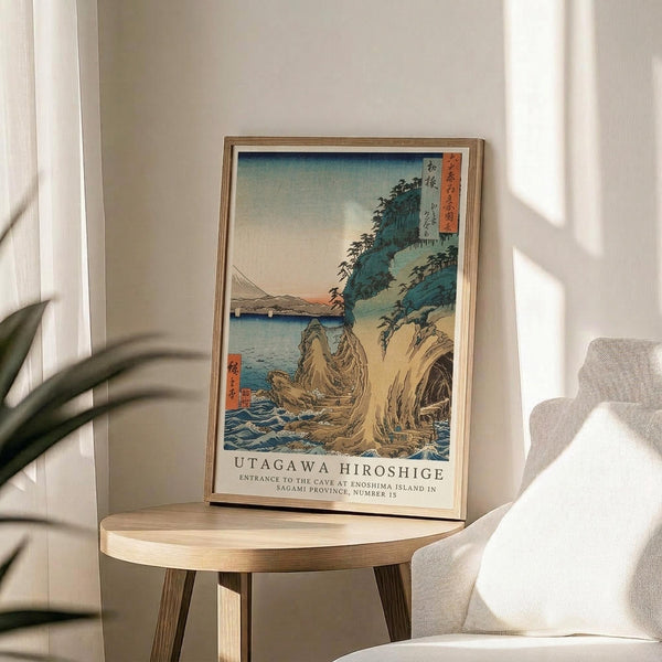 May include: A framed Japanese woodblock print reproduction displayed on a round wooden side table next to a beige upholstered chair. The artwork depicts a coastal landscape with rocky cliffs, blue water, and Mount Fuji in the distance, featuring traditional ukiyo-e style with soft blues, greens, and earth tones. Natural sunlight streams across the minimalist room setting with white walls and a potted plant visible in the corner.