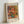 May include: Framed art print of Renoir's impressionist painting showing pink and coral roses in a blue-green vase against warm, colorful brushstrokes. The poster has "VASE OF ROSES" at top and "PIERRE AUGUSTE RENOIR 1890-1900" at bottom on white matting within a light wood frame, leaning against a white wall.