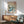 May include: Japanese woodblock print poster in wooden frame showing Mount Fuji, crashing ocean waves in blue and white, pine trees, and flying birds. Displayed above a mid-century modern wooden sideboard with white ceramic lamp. Light green upholstered armchair with wooden frame sits nearby in bright, sunlit room with white walls.