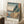 May include: Framed Japanese woodblock print reproduction leaning against white wall, showing coastal scene with rocky cliffs, pine trees, blue water, and Mount Fuji in distance. Warm sunset colors blend from blue sky to peachy horizon. English text identifies the artwork below the image. Small wooden side table with white coffee cup and saucer sits nearby on textured cream rug. Soft natural lighting creates gentle shadows on the wall.