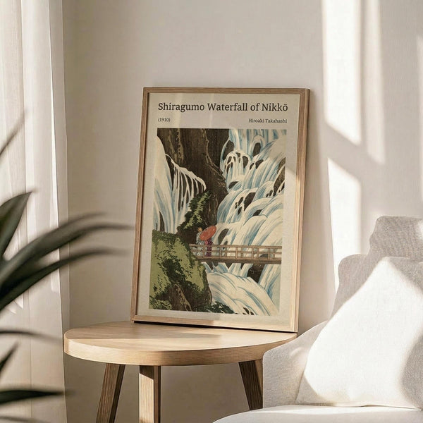 May include: Framed Japanese woodblock print reproduction showing a dramatic waterfall scene with cascading white water, dark rocky cliffs, green vegetation, and small figures on a wooden bridge. The vintage-style artwork is displayed in a light wooden frame on a round wooden side table next to a beige upholstered chair, with a potted plant visible in the corner and natural sunlight streaming across white walls.