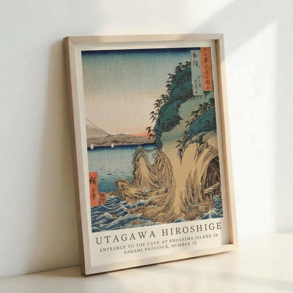 May include: Framed Japanese woodblock print reproduction showing a coastal landscape with dramatic rock formations, crashing waves, and Mount Fuji in the distance. The artwork features traditional ukiyo-e style with blue-green cliffs, turquoise water, and a gradient sky from blue to peachy sunset tones. Red cartouches with Japanese text appear in corners. The print is displayed in a light wooden frame leaning against a white wall, showing the classic Hiroshige composition with detailed wave pat