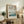 May include: Framed Japanese woodblock print featuring cherry blossoms overhanging a blue river with Mount Fuji in the distance, displayed on a rustic wooden sideboard alongside a fern plant, ceramic vase, and books in a warm, naturally-lit living room setting with neutral tones and vintage furniture.