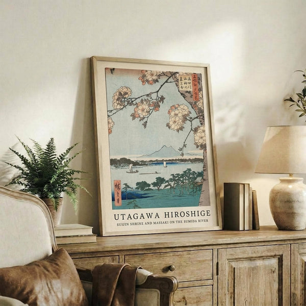 May include: Framed Japanese woodblock print featuring cherry blossoms overhanging a blue river with Mount Fuji in the distance, displayed on a rustic wooden sideboard alongside a fern plant, ceramic vase, and books in a warm, naturally-lit living room setting with neutral tones and vintage furniture.