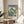 May include: Japanese woodblock print artwork in light wooden frame showing coastal landscape with dramatic rock formations, waterfall, and Mount Fuji in background. Blue water, green hills, and yellow cliffs with cherry blossoms. Text reads "Utagawa Hiroshige Noto Province, Waterfall Bay." Print displayed on round wooden side table next to beige upholstered chair with plant shadows on white wall.