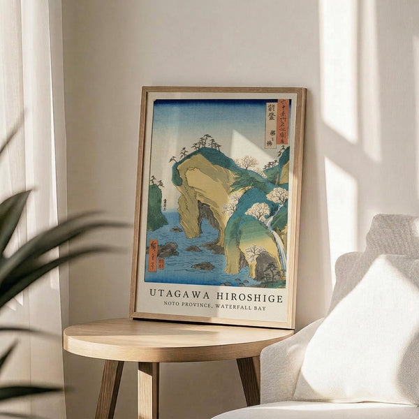 May include: Japanese woodblock print artwork in light wooden frame showing coastal landscape with dramatic rock formations, waterfall, and Mount Fuji in background. Blue water, green hills, and yellow cliffs with cherry blossoms. Text reads "Utagawa Hiroshige Noto Province, Waterfall Bay." Print displayed on round wooden side table next to beige upholstered chair with plant shadows on white wall.
