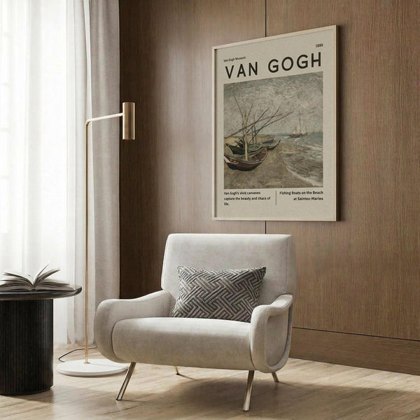 May include: Van Gogh museum poster featuring fishing boats on a beach in muted earth tones, displayed above a beige upholstered armchair with geometric patterned pillow. Modern reading corner with brass floor lamp, dark round side table with open book, and warm wood paneled wall. Natural lighting from window with sheer curtains creates cozy atmosphere.