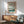 May include: Japanese woodblock print poster in wooden frame hanging above mid-century modern credenza with table lamp. Green upholstered armchair with wooden arms sits nearby. Print shows cherry blossoms framing a serene river scene with Mount Fuji in background, rendered in soft blues and coral tones. Bright natural sunlight creates shadows across white wall and wooden furniture.