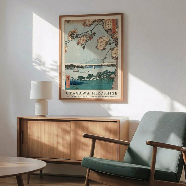 May include: Japanese woodblock print poster in wooden frame hanging above mid-century modern credenza with table lamp. Green upholstered armchair with wooden arms sits nearby. Print shows cherry blossoms framing a serene river scene with Mount Fuji in background, rendered in soft blues and coral tones. Bright natural sunlight creates shadows across white wall and wooden furniture.