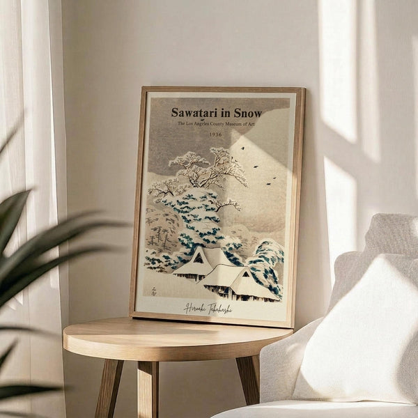 May include: Framed vintage Japanese woodblock print featuring snow-covered houses beneath a large wave and bare tree branches, displayed on a round wooden side table next to a beige upholstered chair. Soft natural lighting creates shadows on the neutral wall behind. The artwork shows muted colors of blue, beige, and white in traditional ukiyo-e style.