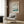 May include: Framed Japanese woodblock print showing cherry blossoms over a river landscape with Mount Fuji in background, displayed on wood-paneled wall above beige upholstered chair with geometric patterned pillow, alongside brass floor lamp and dark side table in modern interior setting