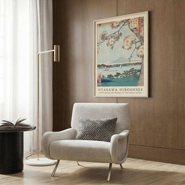 May include: Framed Japanese woodblock print showing cherry blossoms over a river landscape with Mount Fuji in background, displayed on wood-paneled wall above beige upholstered chair with geometric patterned pillow, alongside brass floor lamp and dark side table in modern interior setting