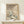 May include: Framed vintage Japanese-style winter landscape poster showing snow-covered traditional houses beneath bare trees and rolling hills in muted beige, blue and white tones. The print sits on a light wood console table next to a small wrapped gift with evergreen sprig. Natural lighting creates soft shadows on cream colored wall.