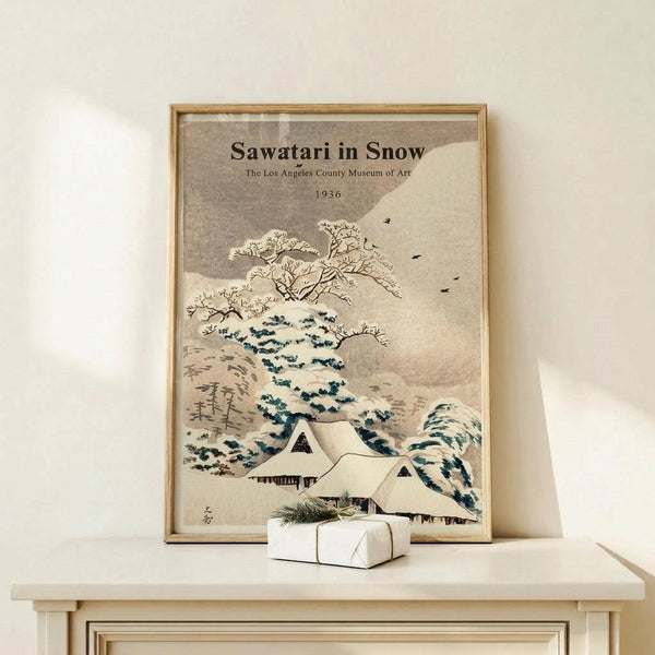 May include: Framed vintage Japanese-style winter landscape poster showing snow-covered traditional houses beneath bare trees and rolling hills in muted beige, blue and white tones. The print sits on a light wood console table next to a small wrapped gift with evergreen sprig. Natural lighting creates soft shadows on cream colored wall.