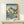 May include: Framed Japanese woodblock print showing coastal landscape with dramatic rock formations, natural arch over blue water, green hillsides with trees, and traditional calligraphy. Print displays in thin wooden frame on light wood surface alongside small white wrapped gift box against cream wall.