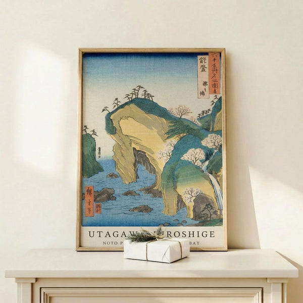 May include: Framed Japanese woodblock print showing coastal landscape with dramatic rock formations, natural arch over blue water, green hillsides with trees, and traditional calligraphy. Print displays in thin wooden frame on light wood surface alongside small white wrapped gift box against cream wall.