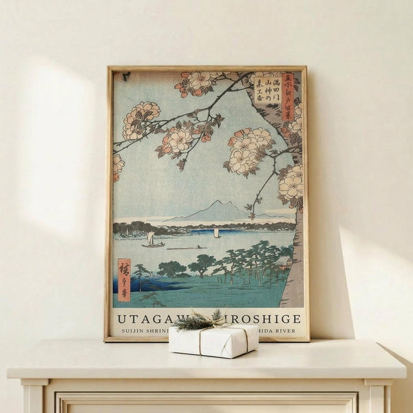 May include: Framed Japanese woodblock print reproduction showing cherry blossoms hanging over a serene river landscape with Mount Fuji in the distance, small boats on blue water, and traditional buildings along the shoreline. The vintage-style artwork features soft pink blossoms, muted blue and green tones, and Japanese text. Displayed on a light wood console table next to a small white wrapped gift with twine and greenery against a cream wall.